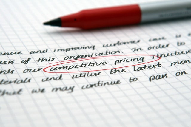 A red pen rests on a notebook filled with handwritten text on squared paper. The phrase competitive pricing is circled in red ink, emphasizing its importance in the document amidst discussions about the effects of globalization.