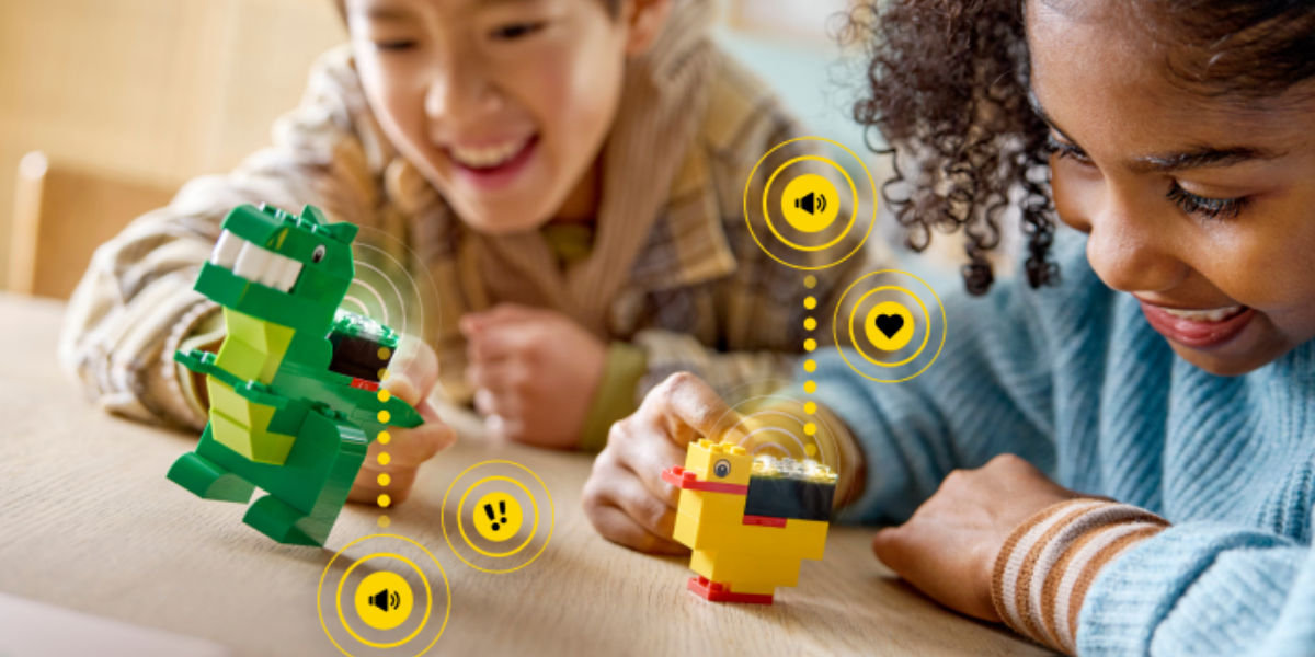 Two smiling children play with colorful LEGO animal figures, a green dinosaur and a yellow duck, on a table. Bright icons and sound waves are illustrated, indicating interactive features.